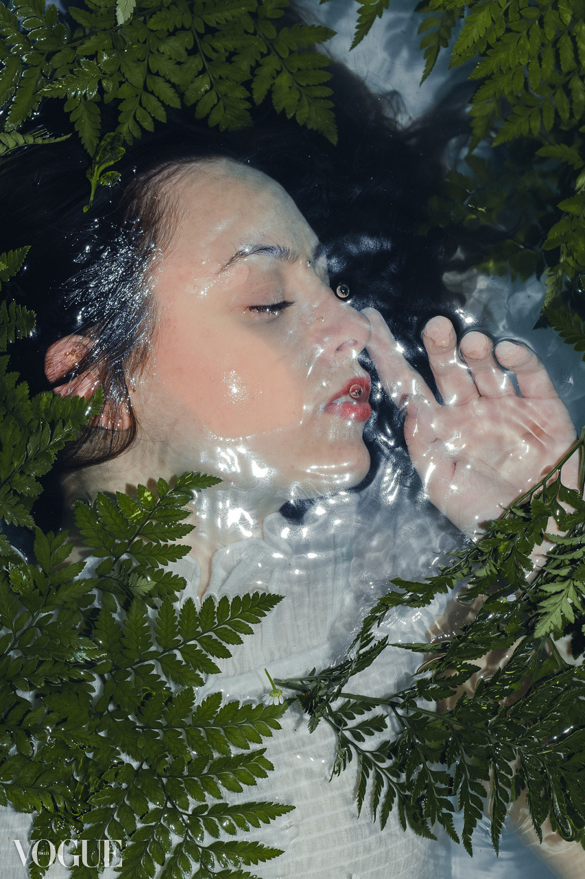 Close view of a face and hand partly submerged in water among wet green fern leaves.