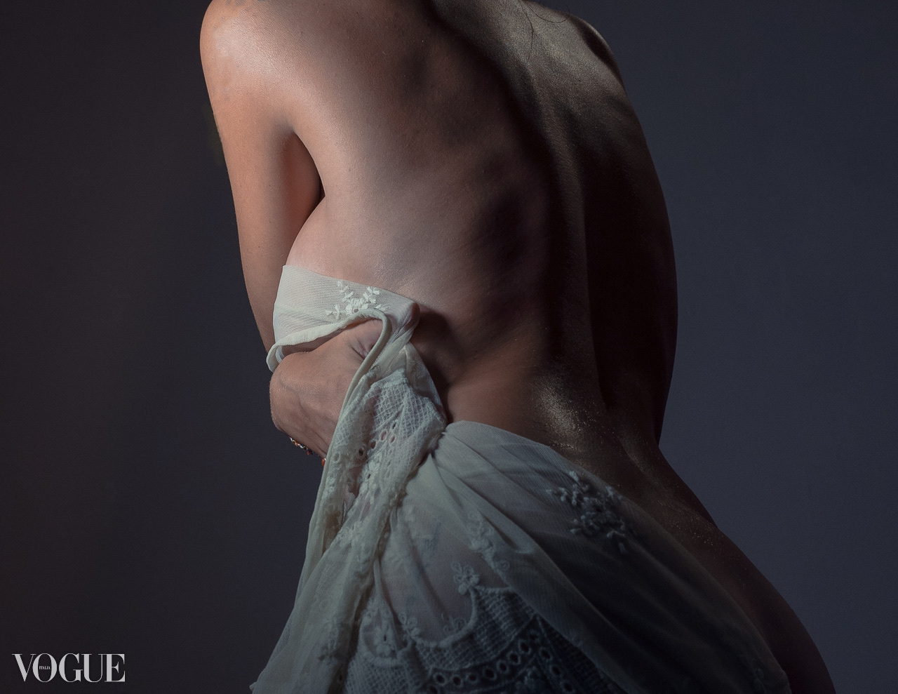 A bare-backed figure gathers pale embroidered fabric against a muted grey background.