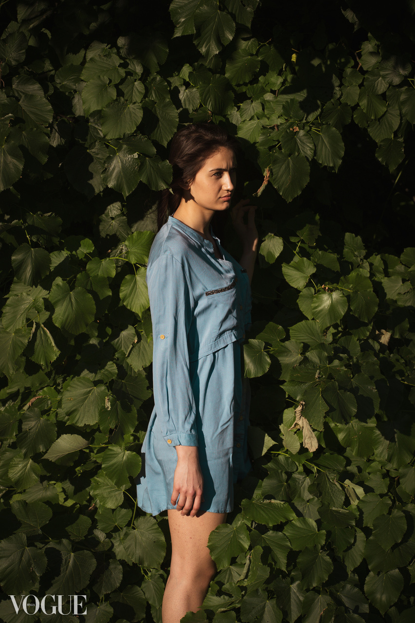 A figure in a pale blue dress stands in profile against dense green leaves in low sunlight.