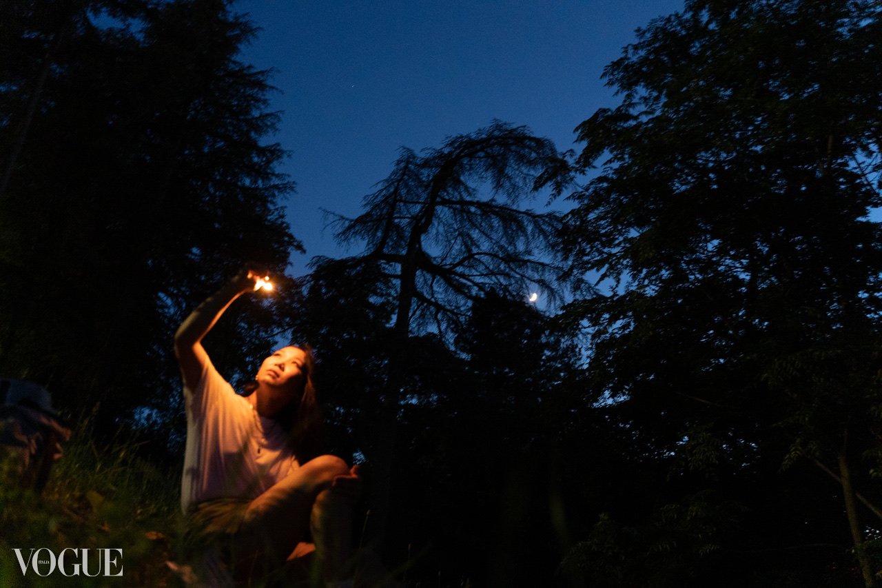 A figure holds a small light above their head in a dark wooded landscape at night.