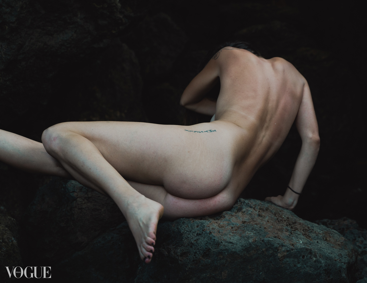 A nude figure folds into a sculptural pose on dark rock against a black background.