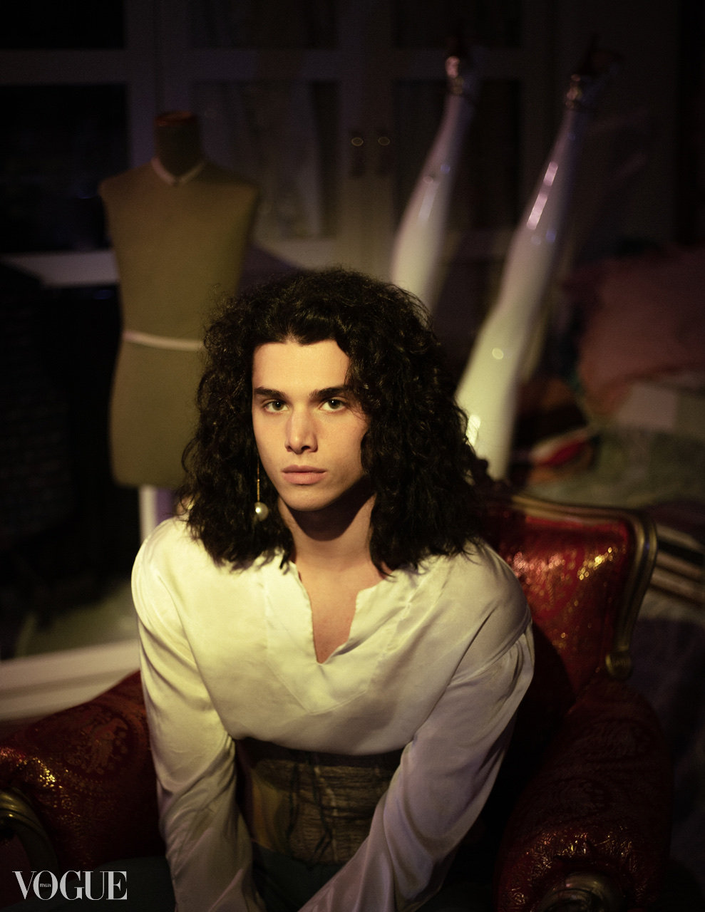 A dark-haired figure in a white shirt sits on a red chair in a dim room with dressmaker's mannequins.