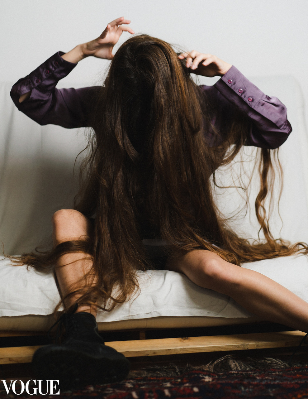 A seated figure in a purple shirt has their face hidden by long brown hair as their legs extend across a white bed.