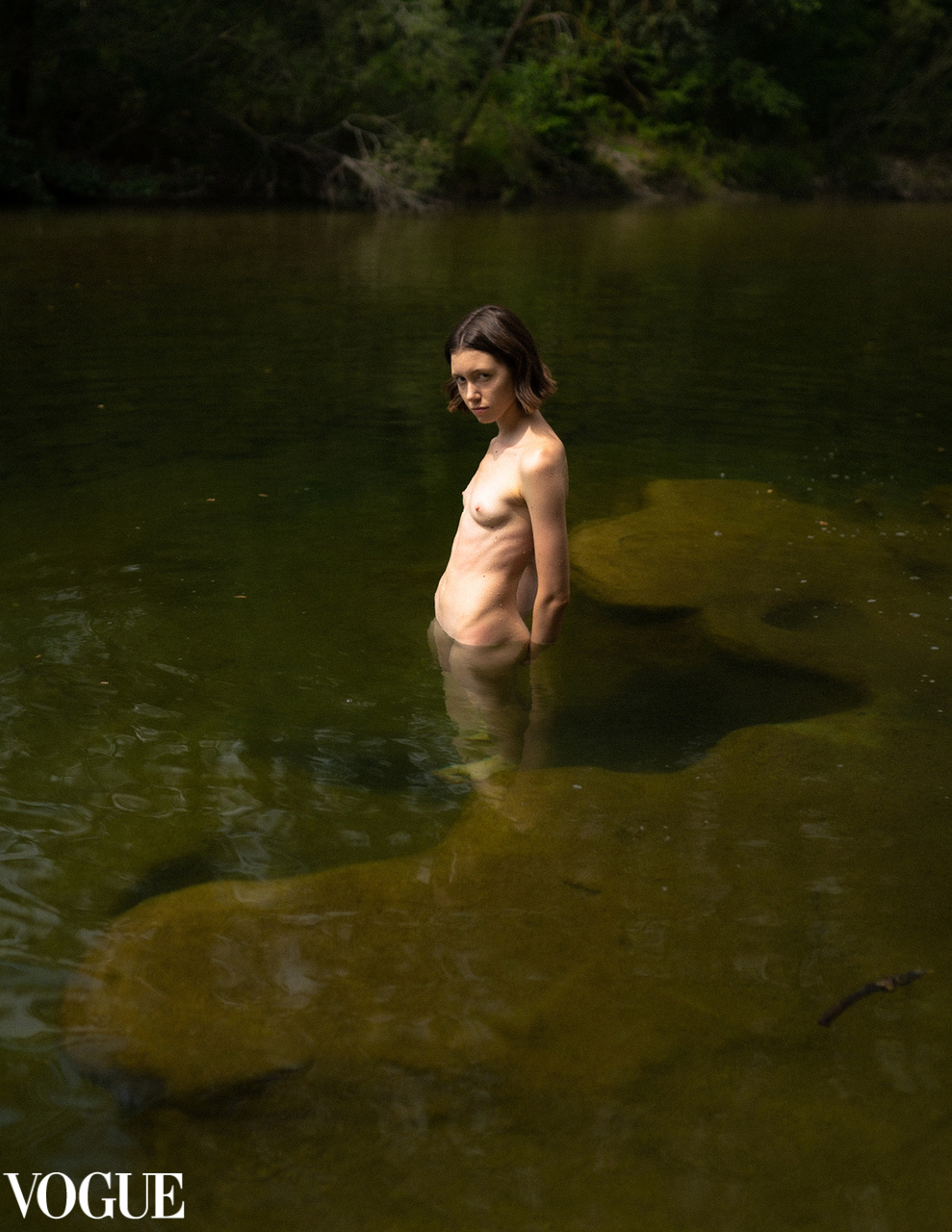 A nude figure stands waist-deep in a dark green river among submerged rocks.