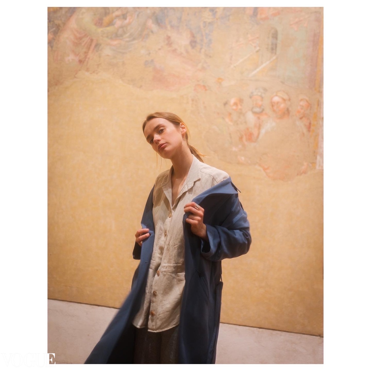 A figure in a blue coat poses before a softly lit wall covered with a faded mural.