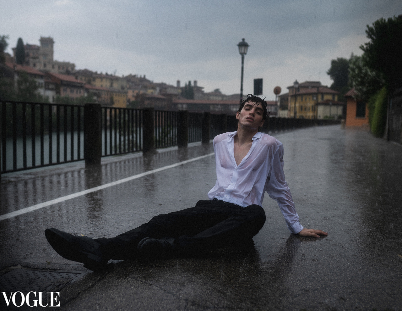 A figure in a white shirt sits on a rain-soaked riverside road beneath a grey sky.