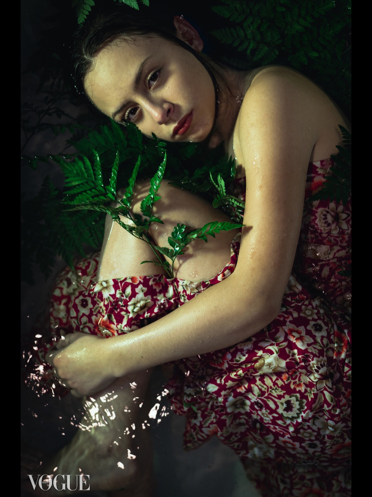 A figure in a red floral dress lies in dark water among wet green ferns and looks toward the camera.